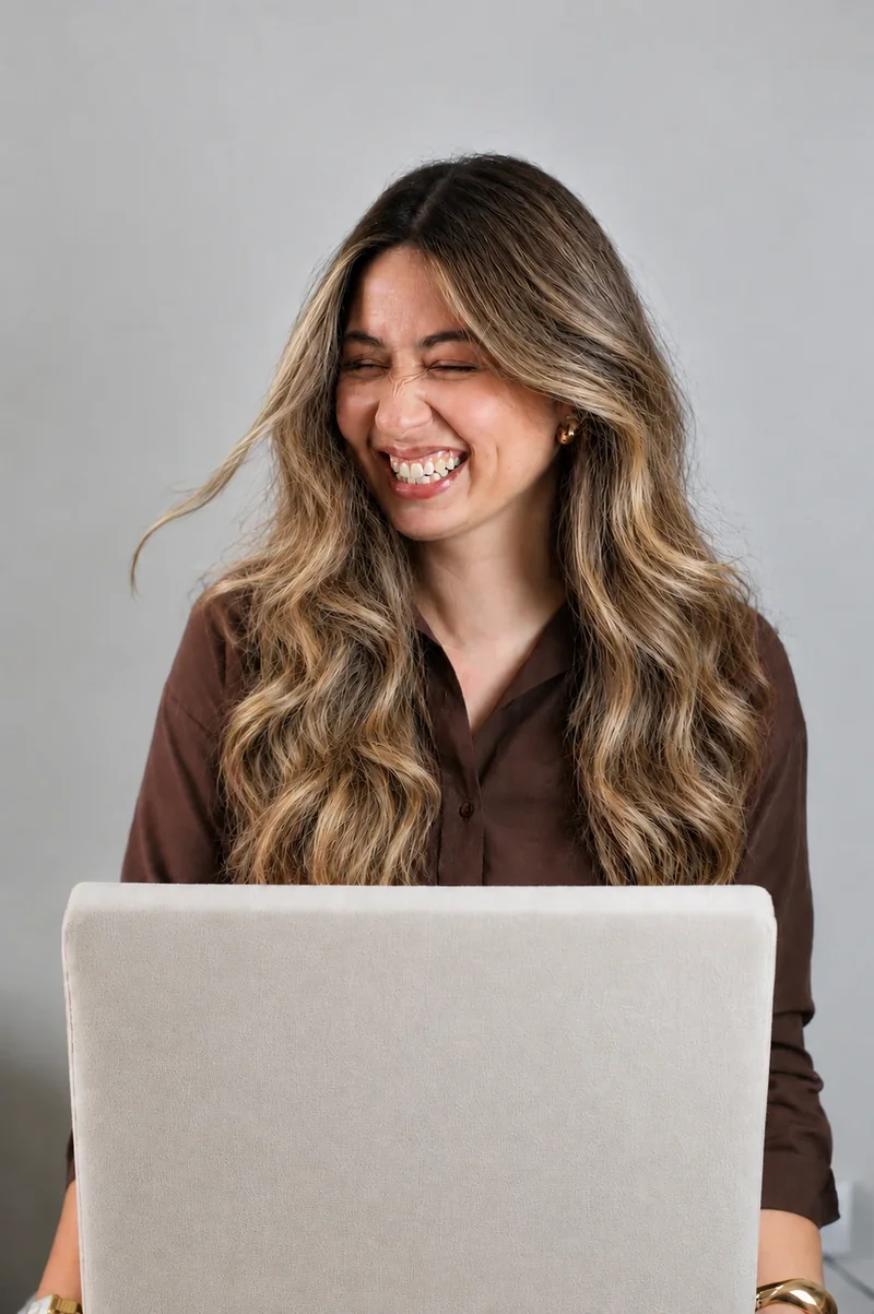 Paloma Ribeiro rindo atrás do notebook, com blusa marrom e acessórios dourados